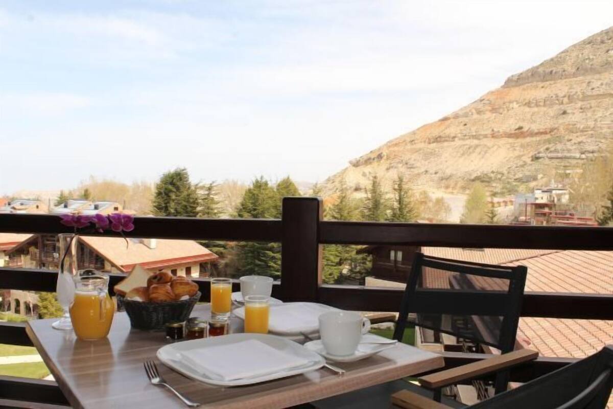 Une table de petit-déjeuner avec des assiettes, des tasses et un panier de croissants, le tout disposé devant un paysage montagneux.