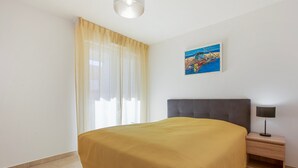 Een slaapkamer met een bed, nachtkastje, lamp, schilderij en een raam met gordijnen.