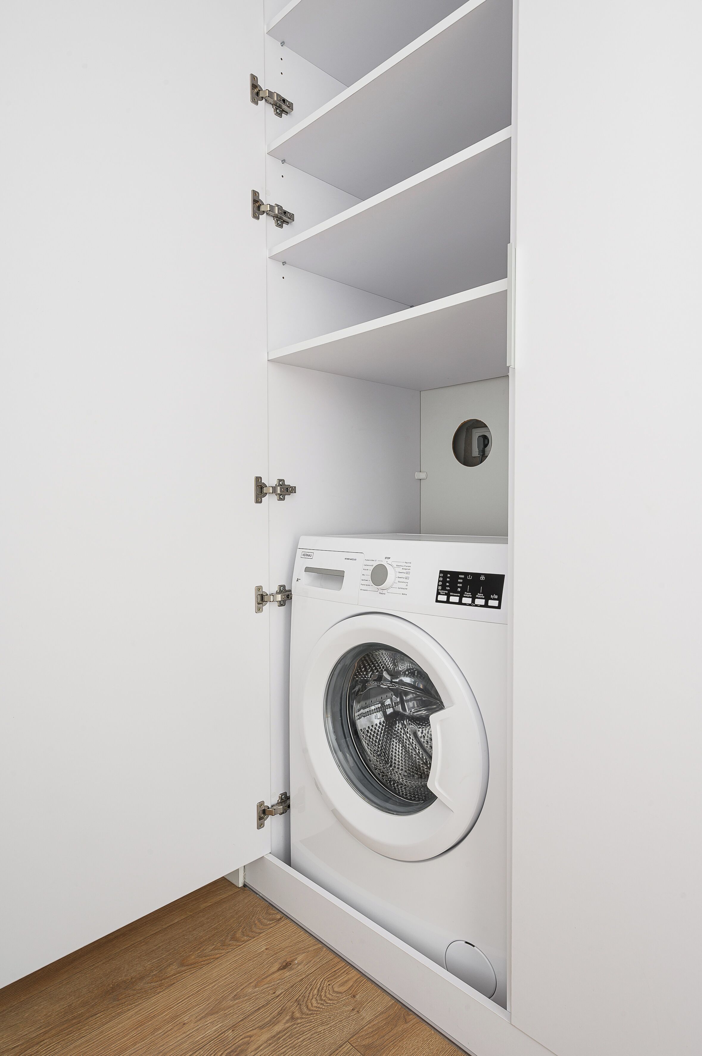 A compact laundry room with a front-loading washing machine, built-in shelving, and a small ventilation unit.