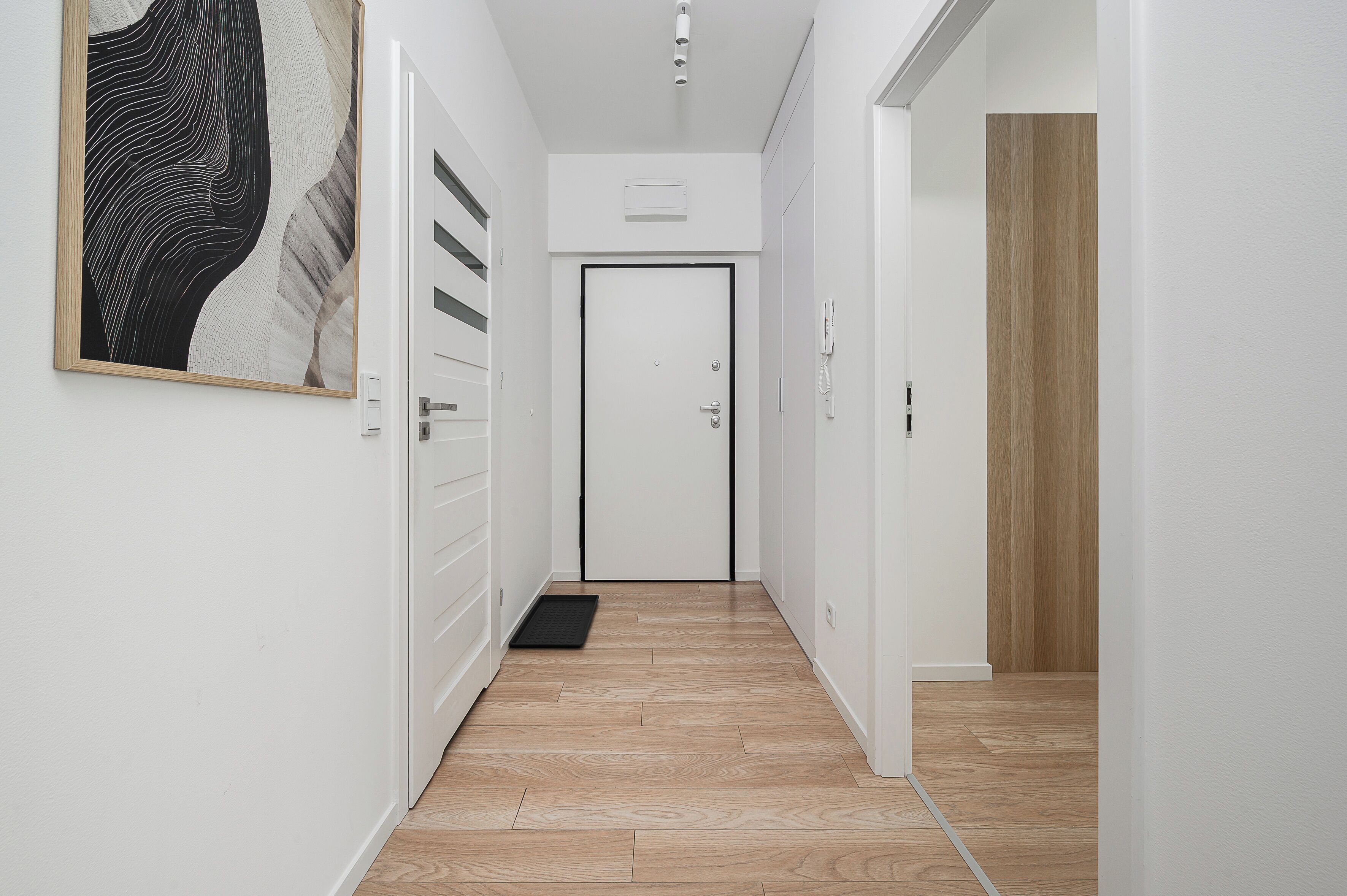 A hallway with a wooden floor, a white door at the end, and a black mat in front of the door.