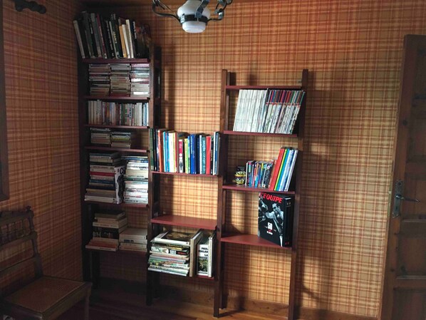 A room with a wooden bookshelf filled with books and DVDs, a small table with a lamp, and a plaid-patterned wall.