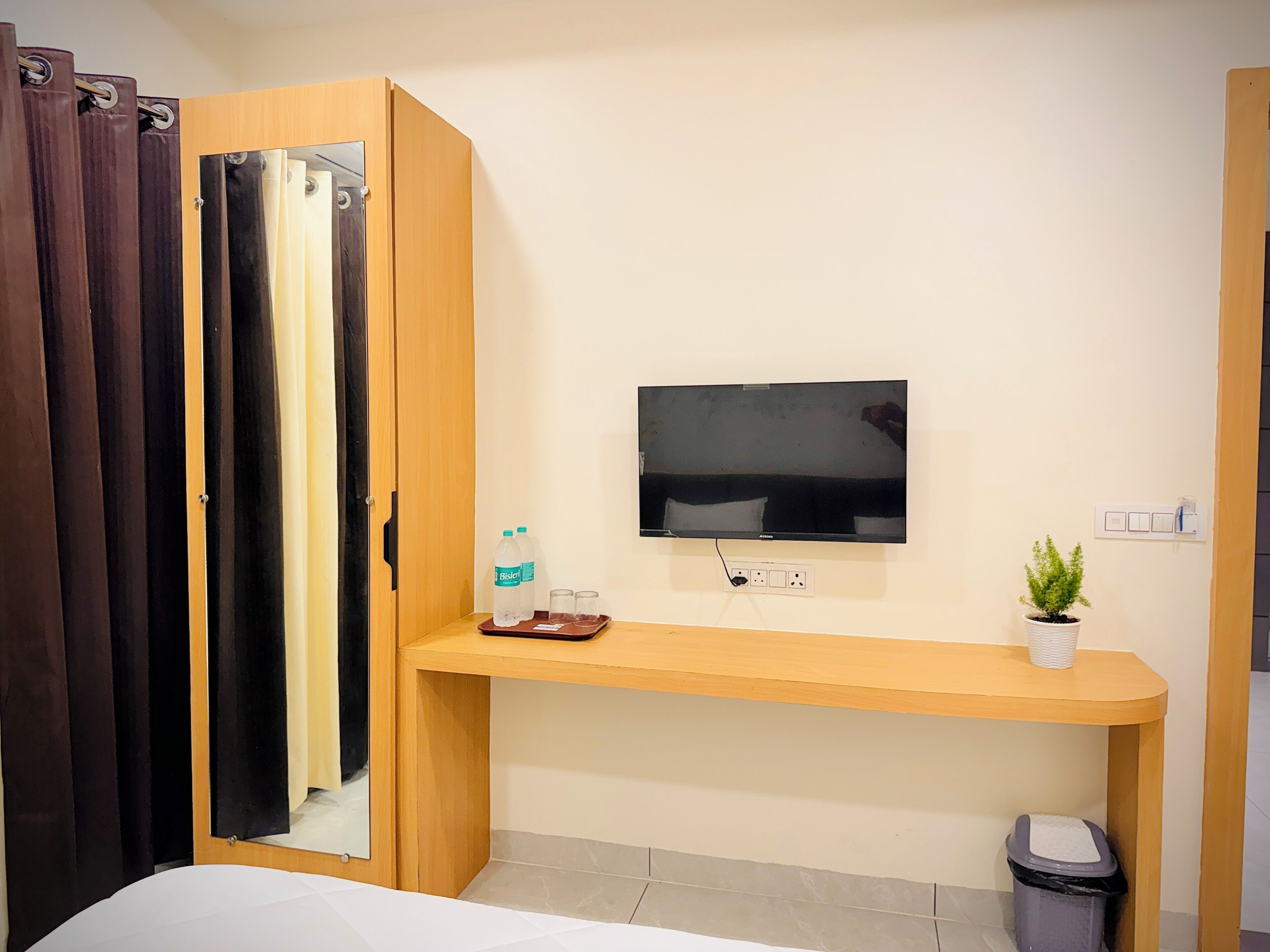 Ein Hotelzimmer mit Holzschreibtisch, einem an der Wand befestigten Fernseher, einer kleinen Topfpflanze und einem Mülleimer.