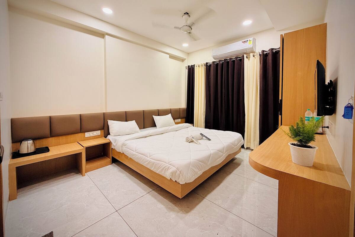 A modern hotel room with a large bed, a bedside table, a small desk, a potted plant, and a mounted TV.