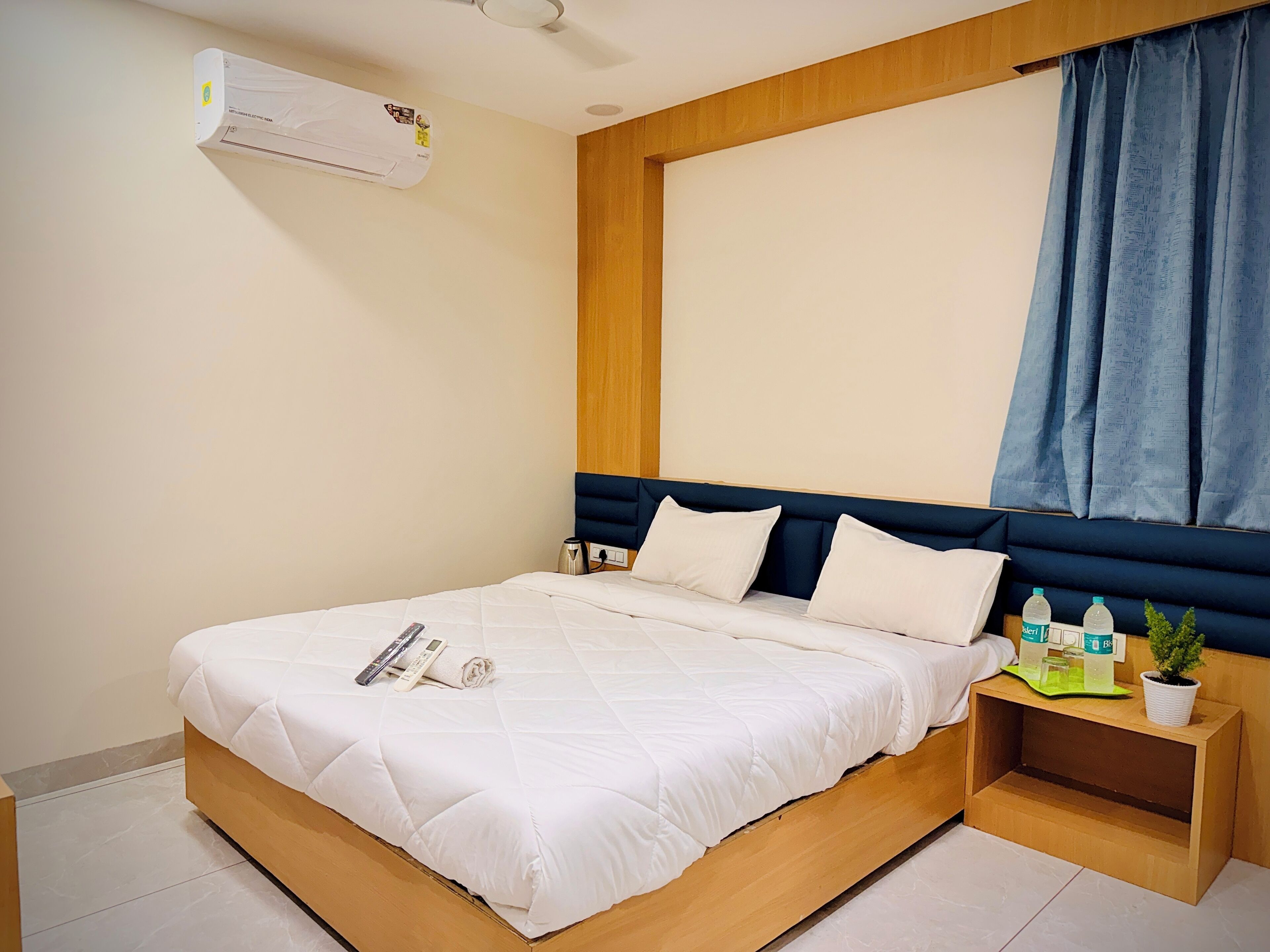 A neatly made bed with a white comforter and pillows, a bedside table with a plant and water bottles, and a wall-mounted air conditioner.
