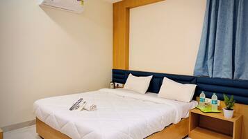A neatly made bed with a white comforter and pillows, a bedside table with a plant and water bottles, and a wall-mounted air conditioner.