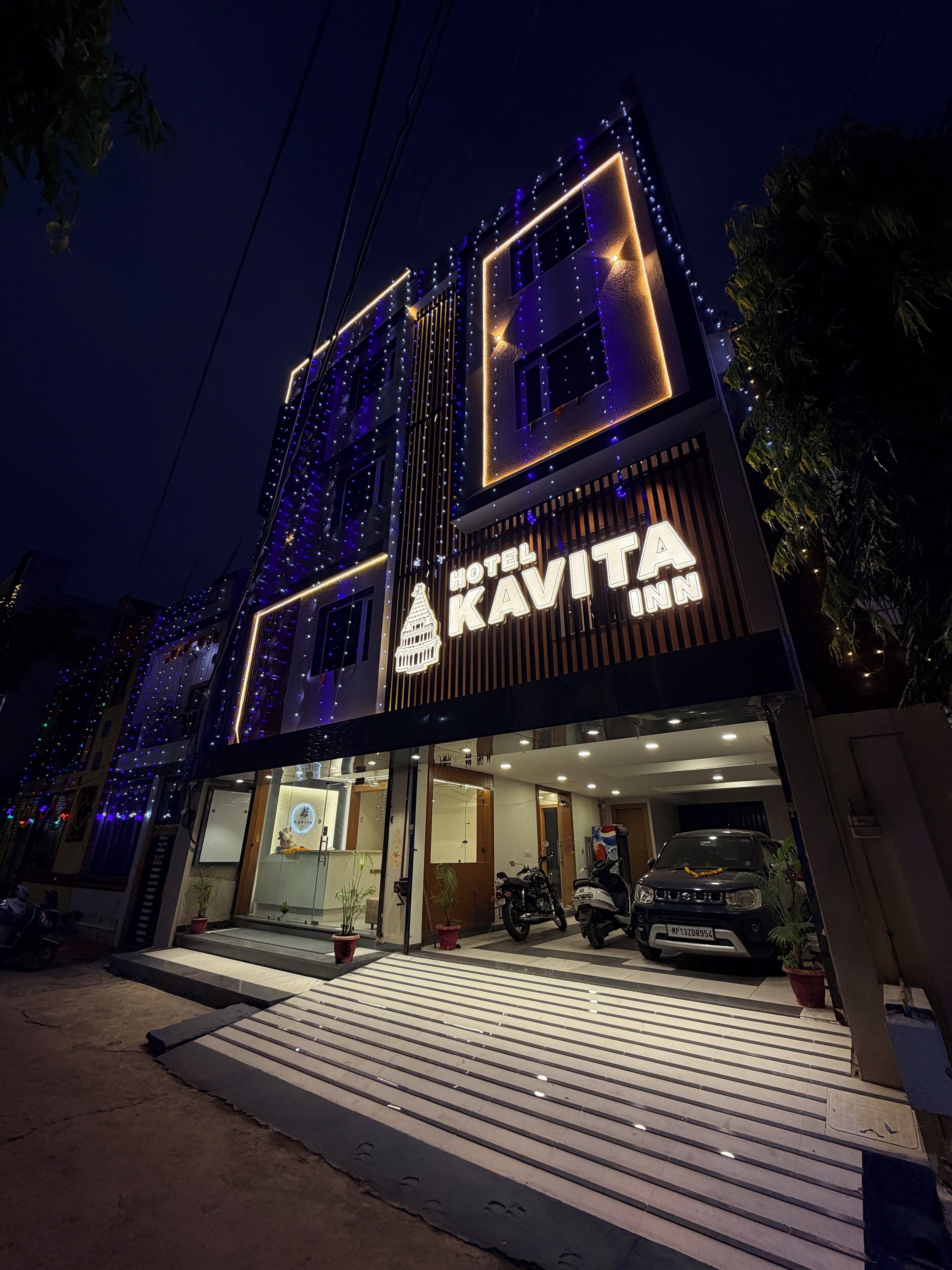 Een hotel genaamd "Hotel Kavita Inn" met een moderne gevel, verlichte signage en een duidelijke ingang, met een motorfiets voor de deur.