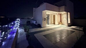 Un edificio moderno con una entrada bien iluminada, un patio con baldosas y vistas al paisaje urbano de fondo.