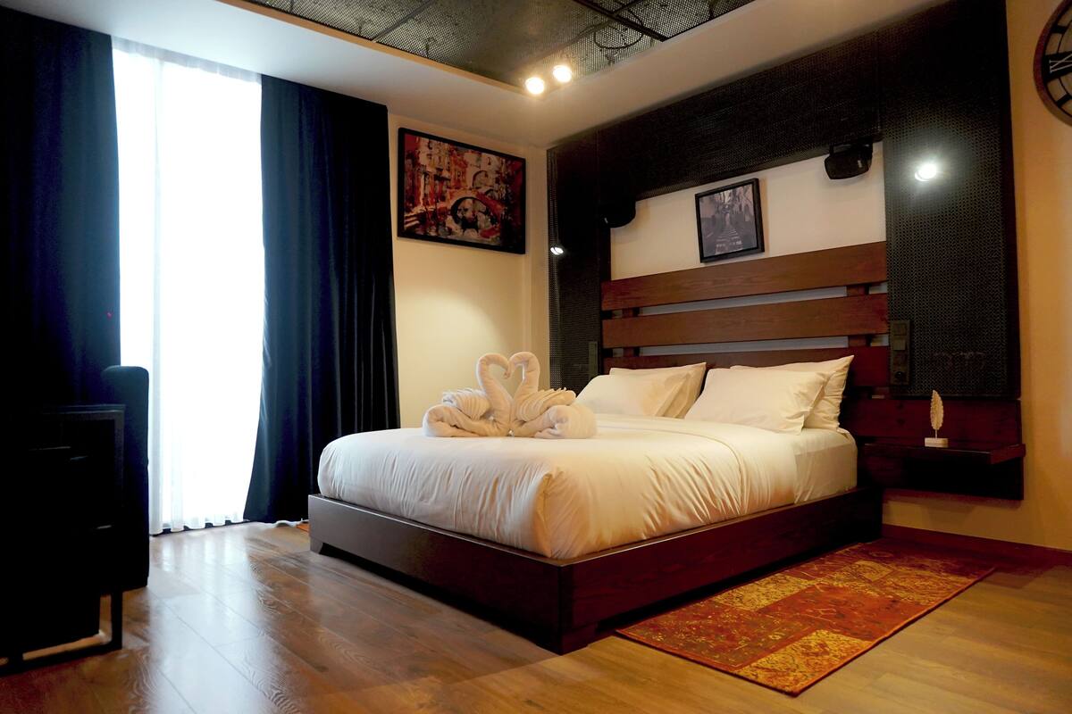 Ein ordentlich gemachtes Bett mit Kopfteil, ein großes Fenster mit Vorhängen und ein Gemälde an der Wand.