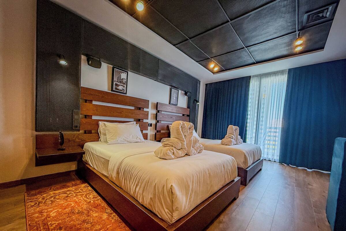 Ein Hotelzimmer mit zwei Betten, einem Holzkopfstück, einem großen Fenster mit Vorhängen und einem gemusterten Teppich.