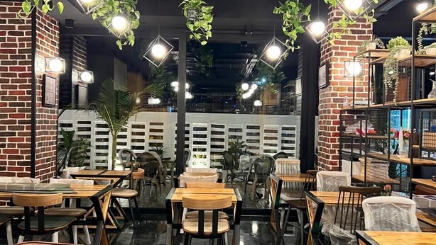 A restaurant interior with brick walls, black and white decor, and hanging plants. Tables and chairs are arranged for dining. - ABM Green (Tangier)