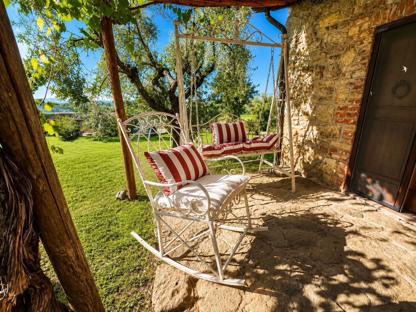 A rustic outdoor seating area with a swing chair and a cushioned bench, a stone wall, and a view of a green landscape.
