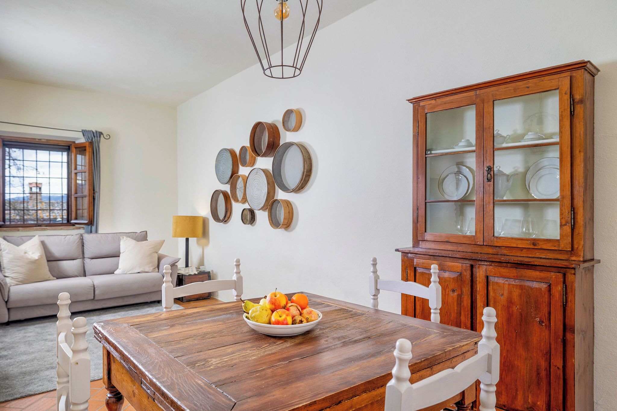 A dining area with a wooden table, white chairs, a wooden cabinet with glass doors, and a wall-mounted fruit bowl.