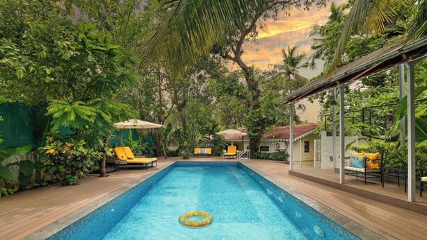 A rectangular swimming pool with a clear blue water surface, surrounded by wooden deck flooring, and shaded by large green trees. - Saligao - Casa De Mello C 1863 (Saligao)