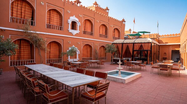 An outdoor dining area with tables and chairs, a fountain, and a building with arches and a flag. - Garrison at Dera Bagseu (Bikaner)