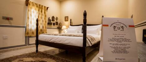 A traditional four-poster bed in a hotel room with a decorative rug, a bedside table, and a lamp. A welcome sign is visible.