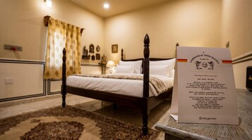 Una cama con dosel tradicional en una habitación de hotel, con una alfombra decorativa, una mesita de noche y una lámpara. Se observa un cartel de bienvenida.