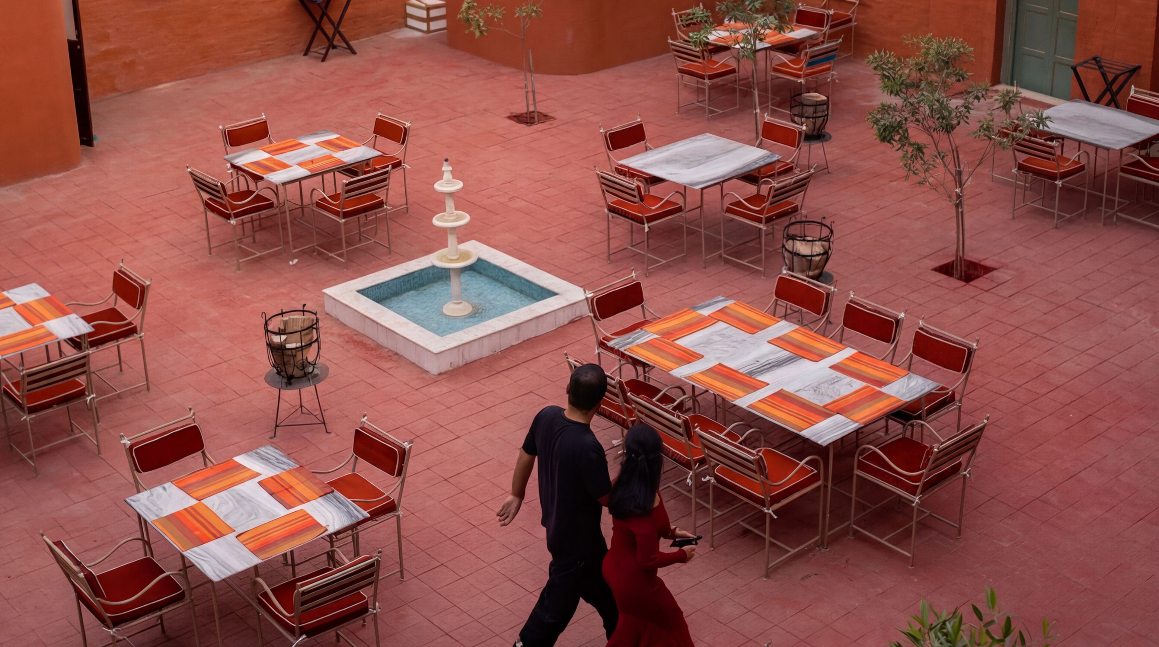Une terrasse extérieure avec plusieurs tables et chaises, une fontaine centrale, et un homme et une femme qui se promènent.