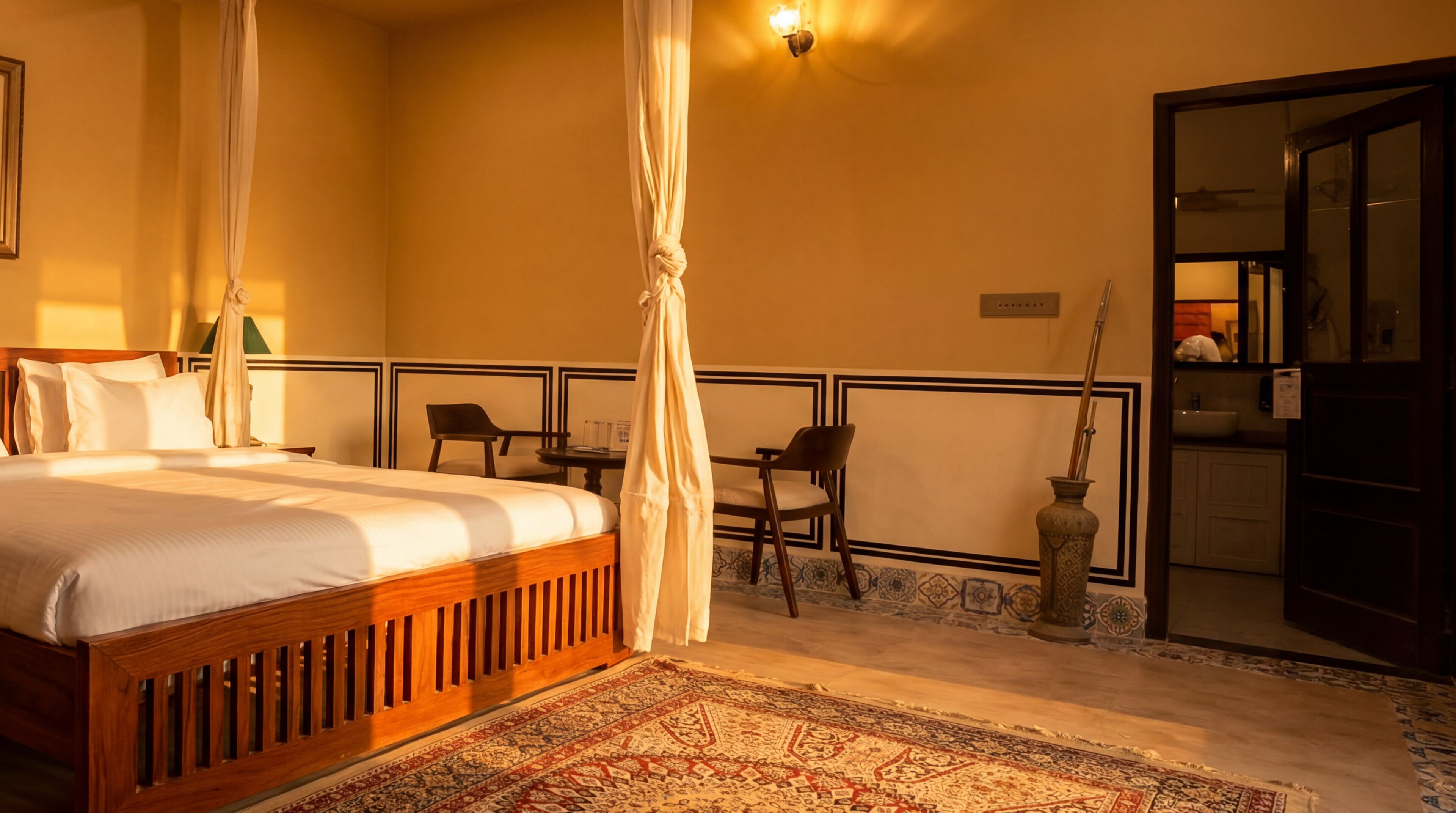 A bedroom with a four-poster bed, a sitting area with chairs, a small table, a decorative vase, and a bathroom visible through an open door.
