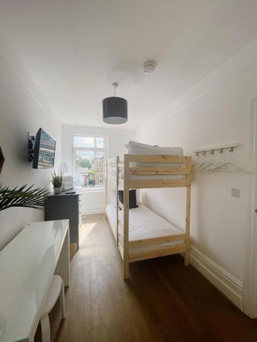 A compact room with a bunk bed, a desk with a chair, a hanging light fixture, and a window providing a view outside.