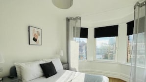 Un dormitorio con cama, mesita de noche, lámpara, ventana con cortinas y vista a la ciudad.