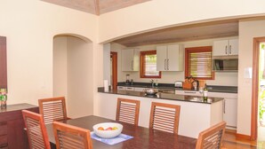 A dining area with a wooden table and chairs, a bowl of fruit, and a kitchen in the background. - Spacious ocean view king bed villa 2 minutes from the restaurant pool and beach (FreeTownst)