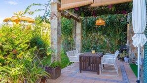 Un patio al aire libre con una mesa de madera y sillas blancas, rodeado de exuberante follaje y una estructura de bambú.