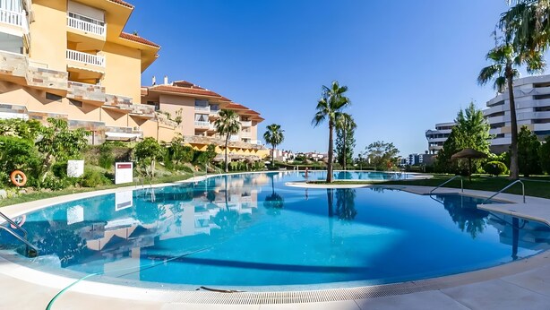 Una piscina rodeada de edificios de varios pisos con balcones, palmeras y un cielo azul despejado.