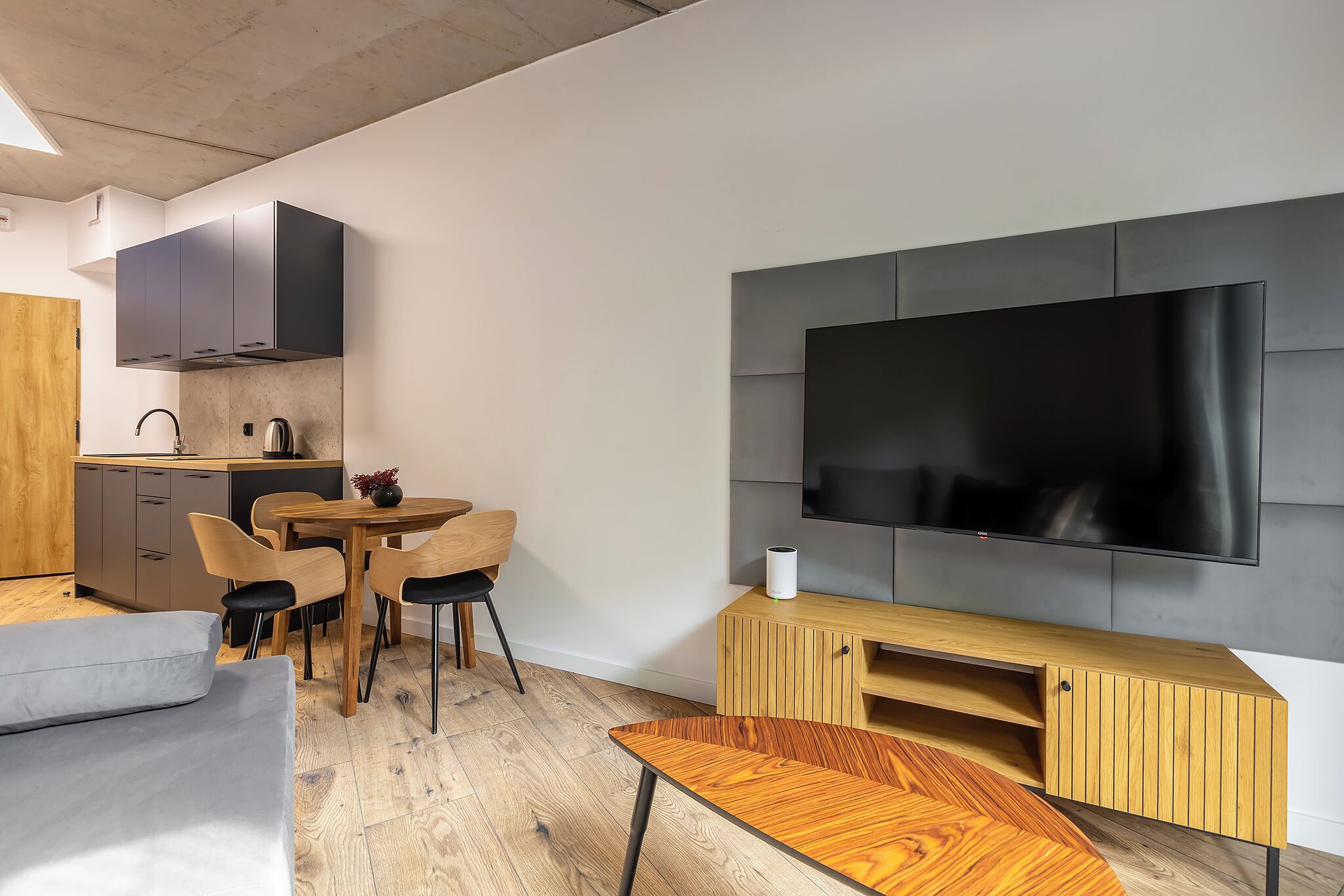 A modern living space with a dining area, a sofa, a TV mounted on a wall, and a kitchen area with cabinets and a sink.