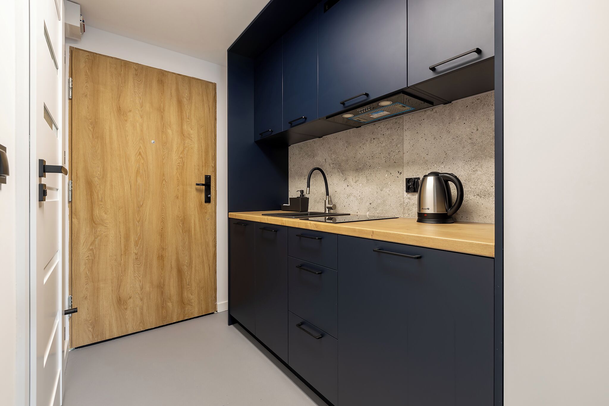 Una cocina moderna con armarios oscuros, una puerta de madera y una tetera de acero inoxidable sobre la encimera.