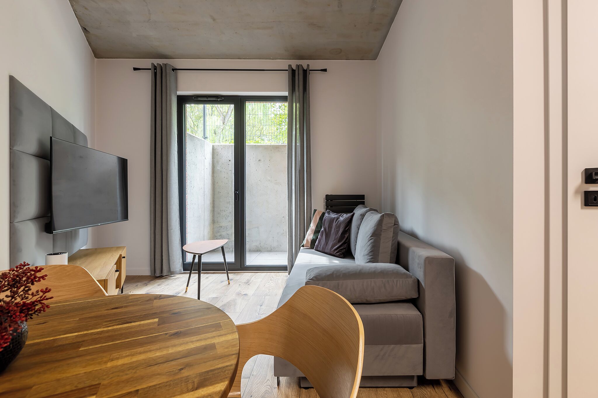 A modern living room with a wooden dining table, a grey sofa, and a flat-screen TV mounted on the wall.