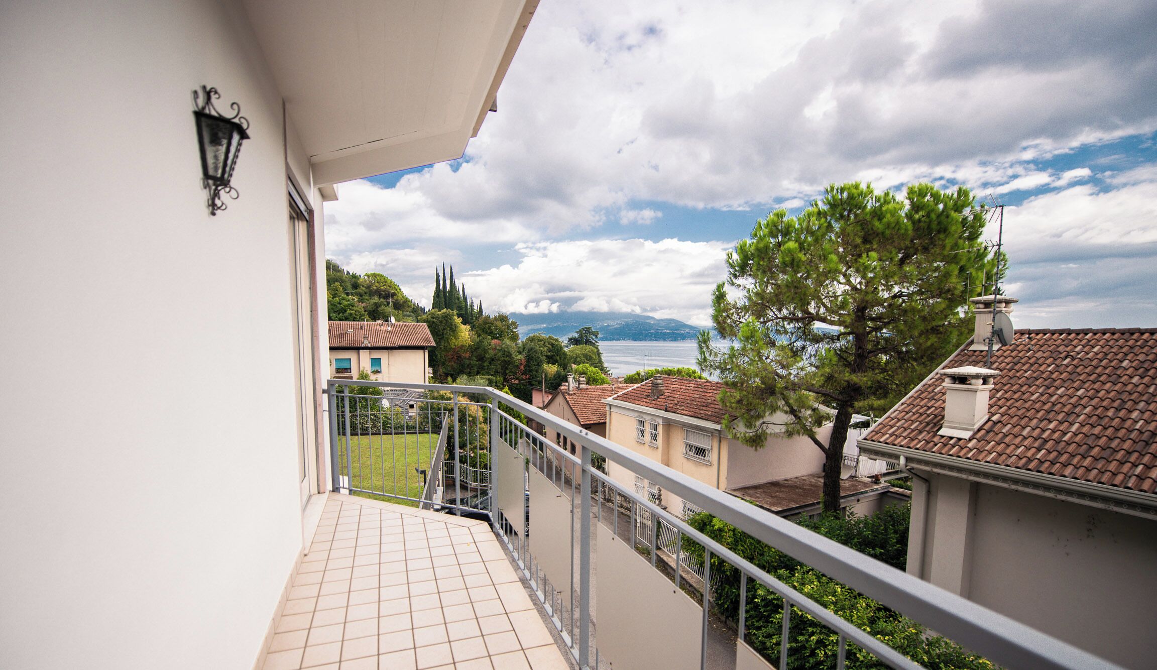 A balcony with a tiled floor, a metal railing, and a view of a lake with buildings and trees in the background.