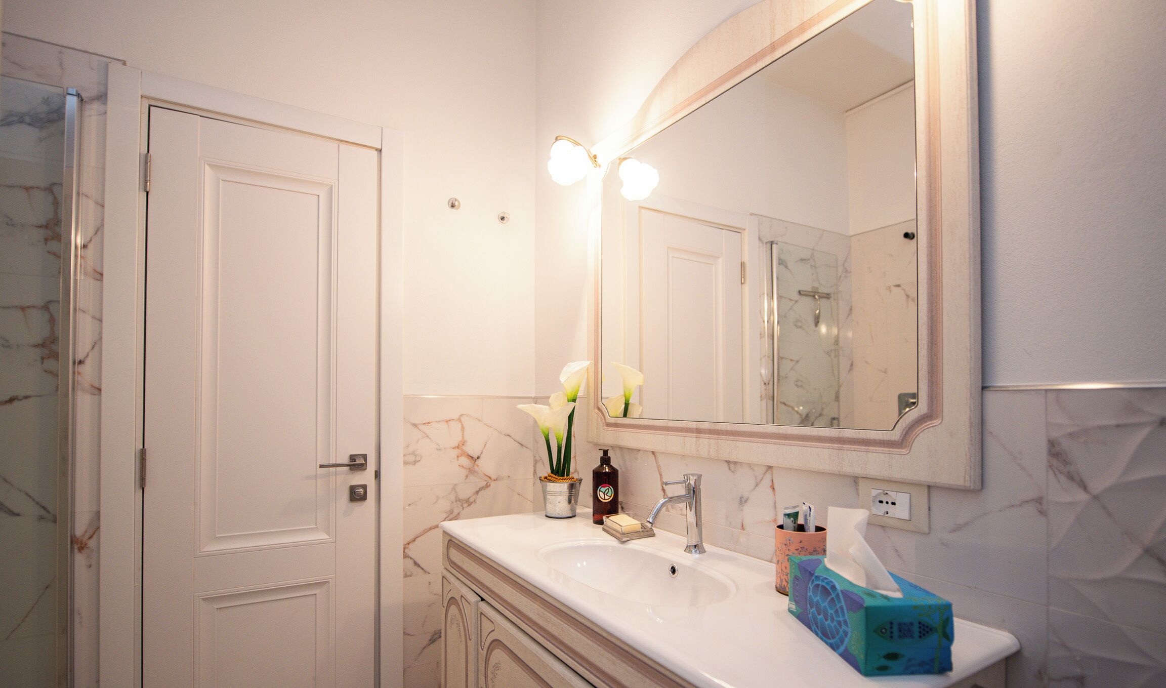 A bathroom with a marble sink, a large mirror with lights, a door, and a shower area.