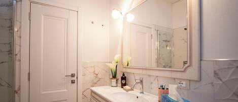 A bathroom with a marble sink, a large mirror with lights, a door, and a shower area.