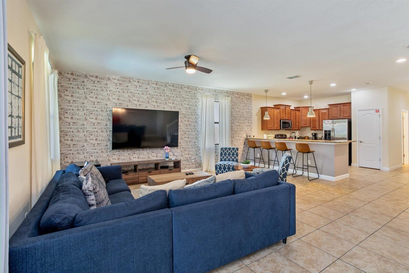A modern living room with a large flat-screen TV, a blue sofa, and a kitchen area with wooden cabinets and a bar stool.
