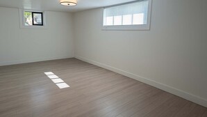 Una habitación con piso de madera, paredes blancas, un ventanal pequeño y una lámpara en el techo.