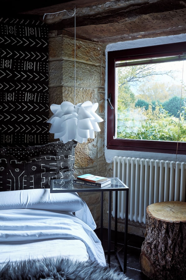 A bedroom with a stone wall, a window with a view of greenery, a bed with patterned pillows, and a wooden stump.