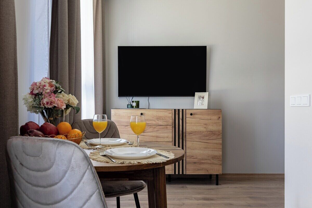 Un coin repas avec une table ronde dressée pour deux personnes, un bouquet de fleurs et un saladier de fruits. Un téléviseur à écran plat est fixé au mur, au-dessus de la table.