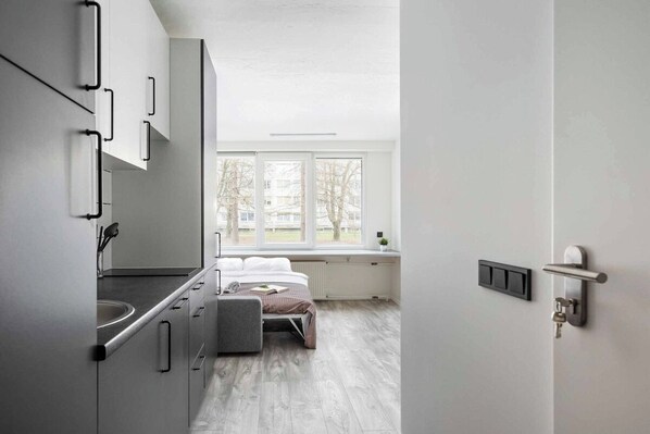 A modern kitchen with a built-in stove, a window with a view of trees, and a bed with a gray headboard in the adjacent bedroom. - River to the City Self check in (Vilnius)