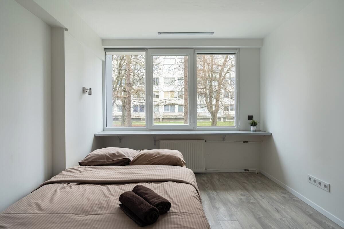 A bedroom with a large window, a radiator, and a bed with a rolled-up blanket.