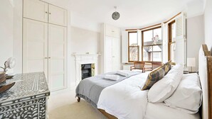 A bedroom with a bed, a dresser, a fireplace, and a window with shutters. - Charming Two-Bedroom Home in London (Greater London)