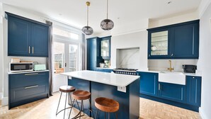 A modern kitchen with dark blue cabinets, a white island with stools, a microwave, and a gas stove. - Charming Two-Bedroom Home in London (Greater London)
