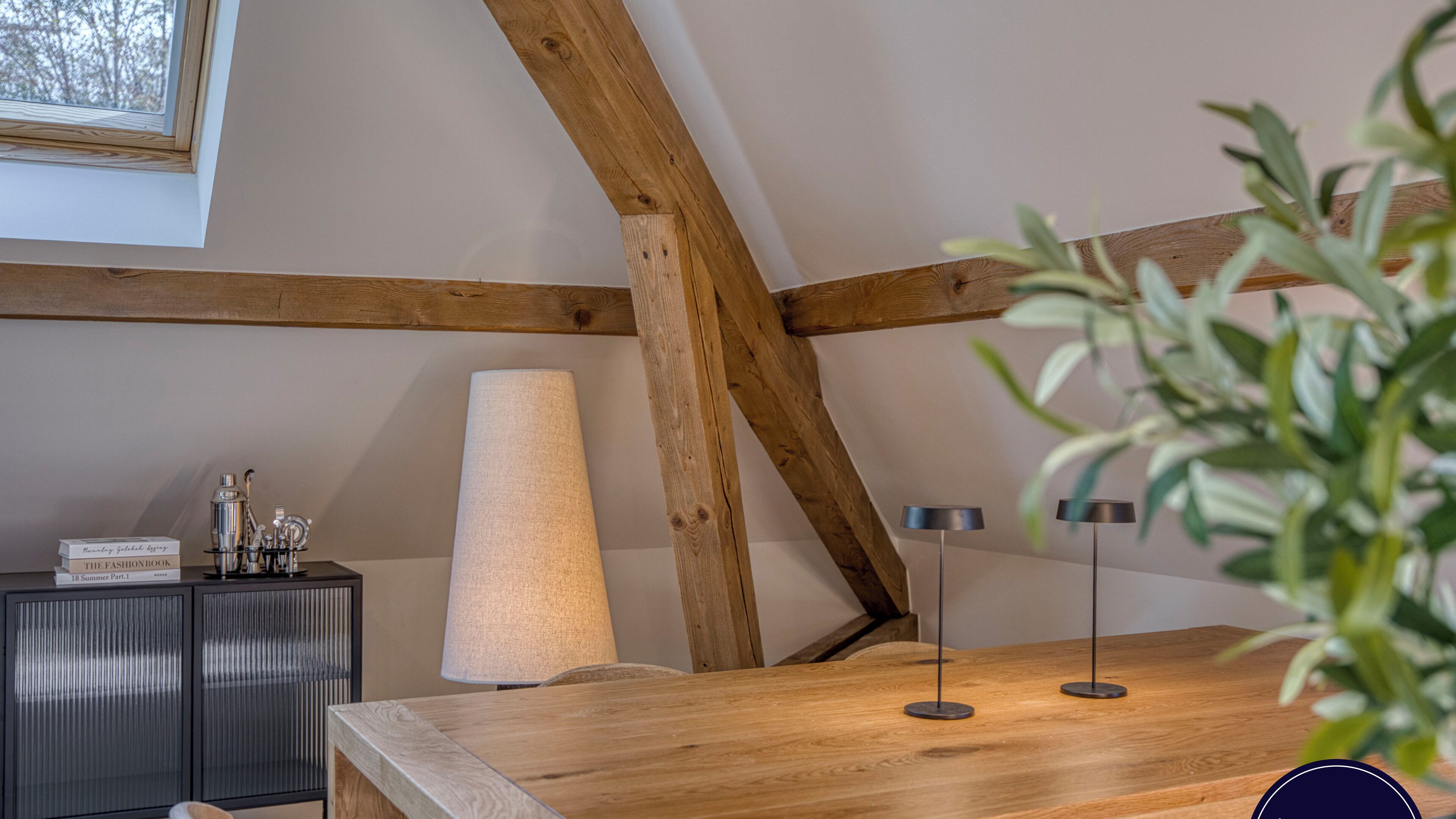 Une table en bois avec une lampe, un petit meuble et une plante, dans une pièce dont les poutres en bois sont apparentes et qui possède une fenêtre.