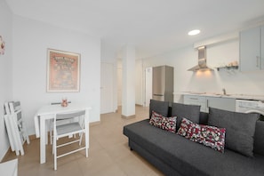 A modern living room with a grey sofa, a white dining table, and a kitchen area with white cabinets and a stainless steel range hood.