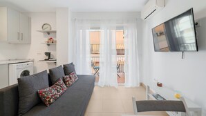 Ein modernes Wohnzimmer mit grauem Sofa, Flachbildfernseher, Waschmaschine und einem Balkon mit Stuhl und Tisch.