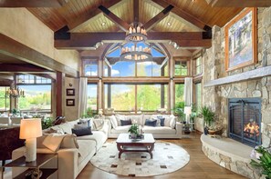 A spacious living room with a fireplace, large windows, and a chandelier. - Private Estate 🏔️ 9,400 sq ft ✨ Game Room 🎱 Steam Sauna 💦 1.5 Acres 🧖‍♀️ Spa (Silverthorne)