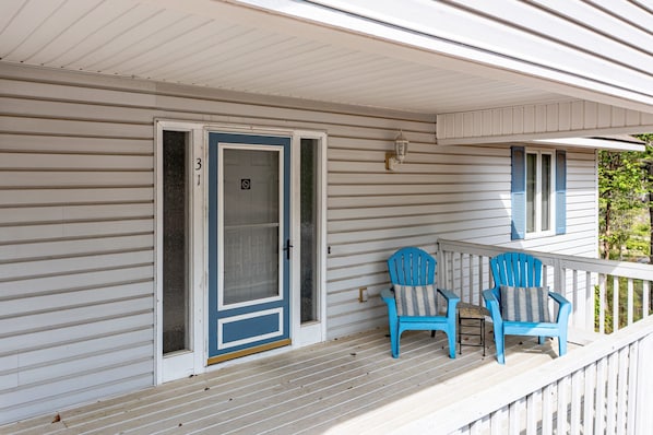 A covered porch with a sliding glass door, two blue Adirondack chairs, and a small table with a striped cushion.