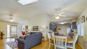 A spacious living room with a gray sofa, wooden dining table, and chairs. There is a kitchen area with cabinets and a refrigerator.