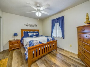 Une chambre à coucher avec un lit en bois, une table de chevet, une commode, un ventilateur de plafond et une fenêtre avec des rideaux bleus.