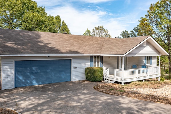 A single-story house with a blue garage door, a covered porch with a white railing, and a small green bush in front.