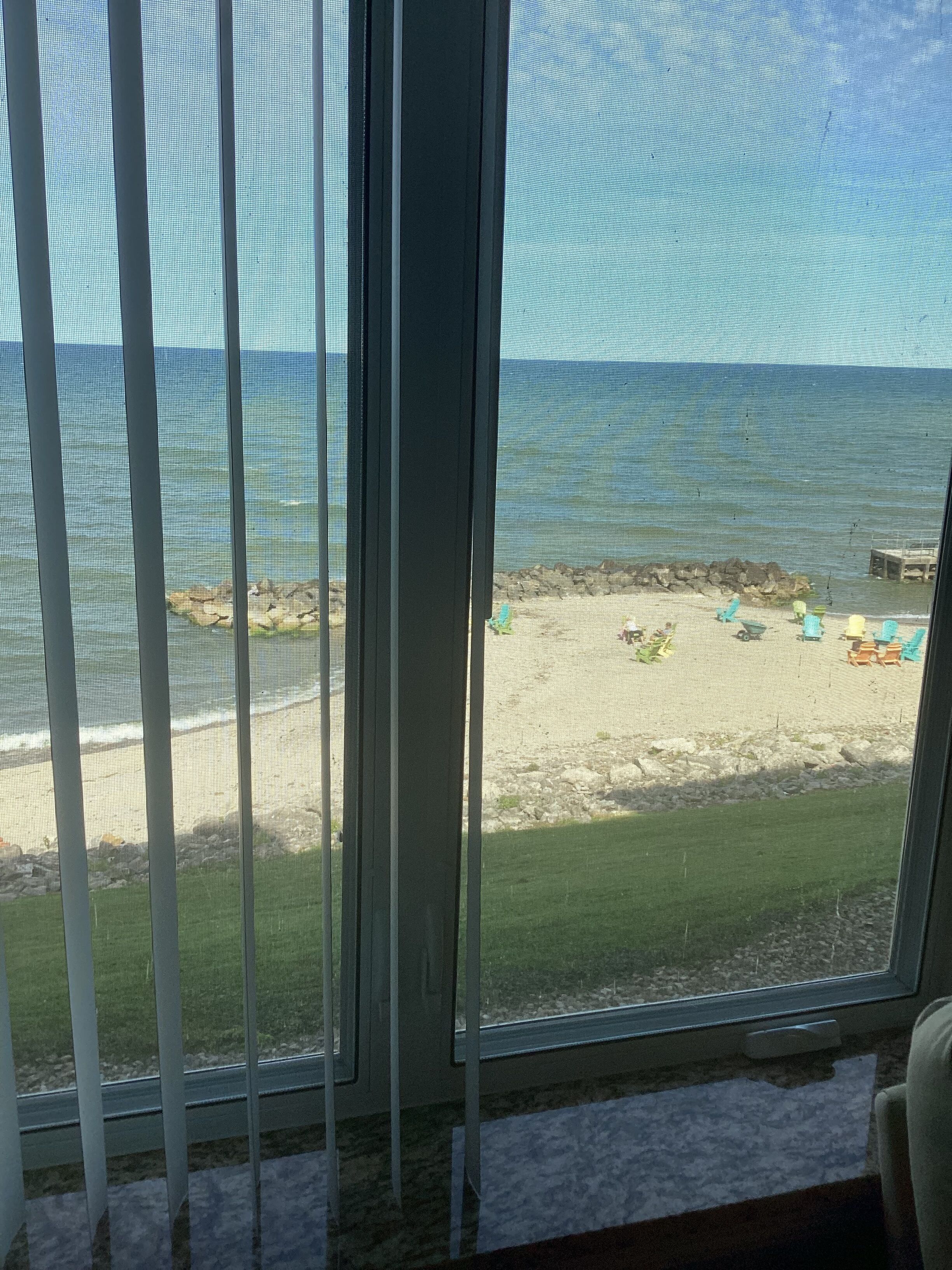A view through a window with vertical blinds, overlooking a beach with a rocky shoreline and a few scattered chairs.